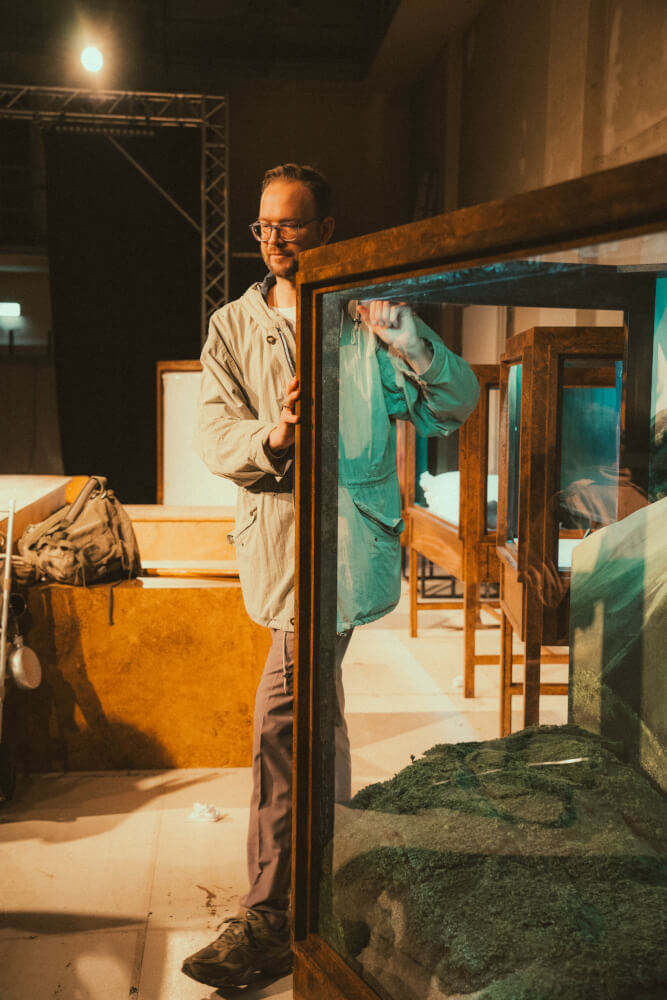 „Die Reise zum Mittelpunkt der Erde“ am Staatstheater Augsburg. Ein Probenfoto mit Wiard Witholt, der einen Rahmen einer Exponatenvitrine festhält.