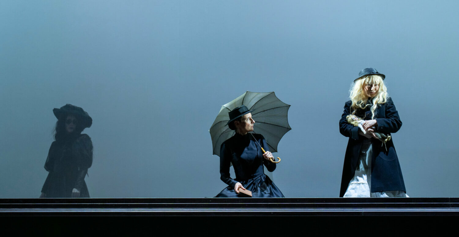 Drei Personen auf einer Bühne, schwarz-englisch-viktorianisch gekleidet, eine mit einem Schirm scheinbar in starkem Wind, einem Sturm sitzend
