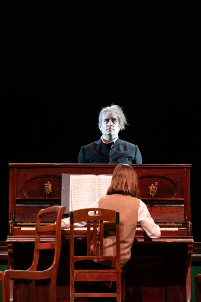 ein Junge am Klavier mit dem Rücken zum Publikum, dahinter geisterhaft ein Mann, der stumm ins Publikum spricht