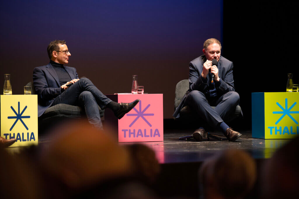 Zu sehen sind zwei Personen sitzend nebeneinander auf der Bühne.