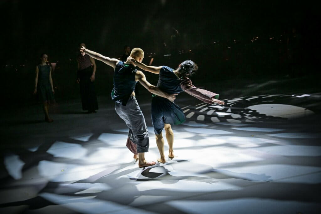 Zu sehen sind zwei Tänzer:innen, die sich aneinander festhalten und drehen auf Reflexionen von Licht.