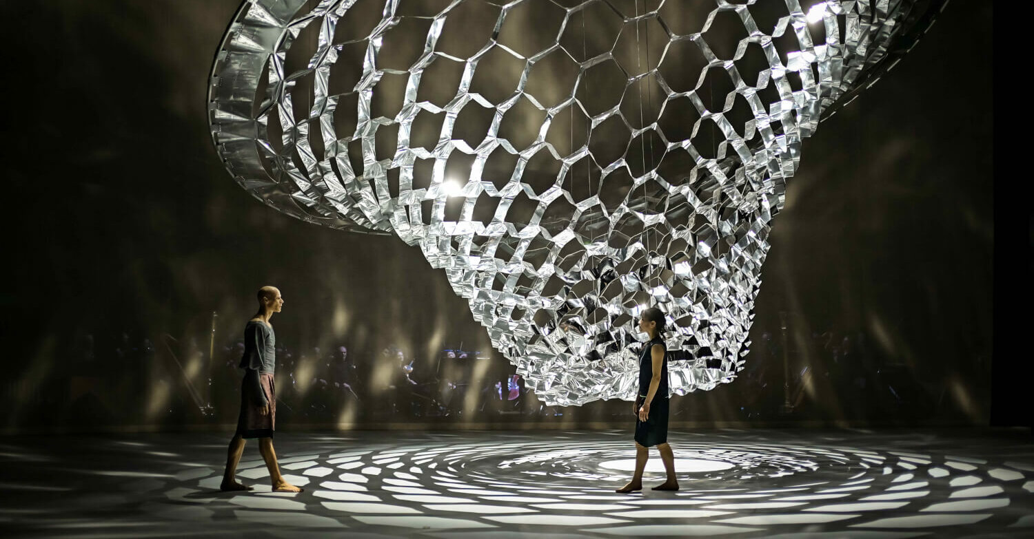 Zu sehen sind zwei Tänzer:innen des Hessischen Staatsballetts unter einer großen, reflektierenden Skulptur.