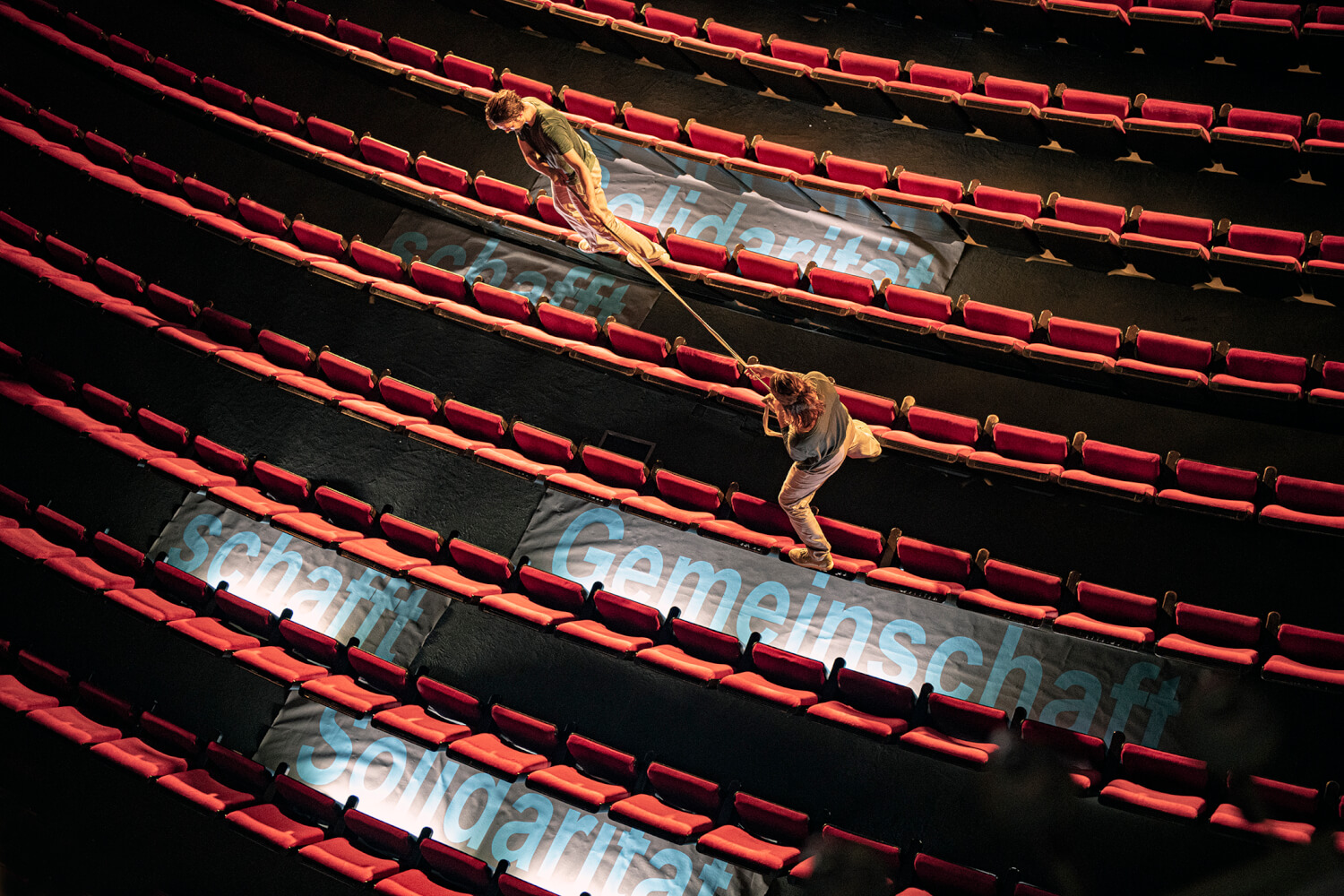 Szene aus Gregor Zölligs choreografischem Parcours „Die Zeit ist reif – Ein Manifest für die Gesellschaft“ am Staatstheater Braunschweig. In einem leeren, rotbestuhlten Zuschauerraum stehen zwei Menschen auf den Rückenlehnen der Stühle, zwei Reihen auseinander und ziehen an einem Seil wie beim Tauziehen. Auf dem Boden zwischen den Stuhlreihen steht "Solidarität schafft Gemeinschaft schafft Solidarität.