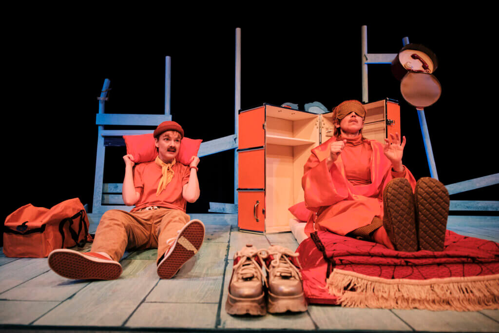 Sylvana Schneider und Jana Alex Rödiger sitzen auf dem Bühnenboden. Beide sind komplett orange, mit Kleidung und Körper. Rechts sind diverse Gegenstände aufgetürmt, auch alle in orange.