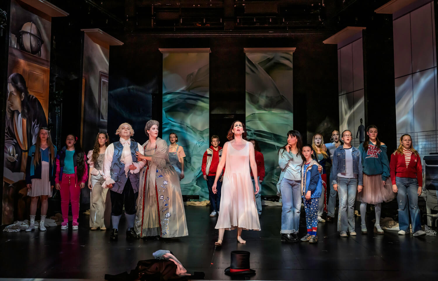 Aufführungsfoto von „Homo Oeconomicus“ von Andrea Tarrodi an der Staatsoper Hannover. Menschen verschiedenen Alters stehen nebeneinander auf der Bühne, in der Mitte eine Frau barfuß mit einem weißen, leichten Kleid.