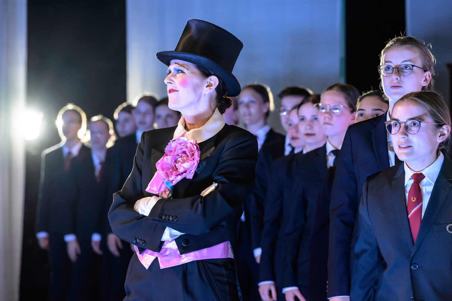 Aufführungsfoto von „Homo Oeconomicus“ von Andrea Tarrodi an der Staatsoper Hannover. Kinder in dunklen Anzügen und roter Krawatte und weißem Hemd stehen in einer Reihe. Davor eine Frau im Frack mit Zylinder, die die Arme verschränkt und verkniffen guckt.