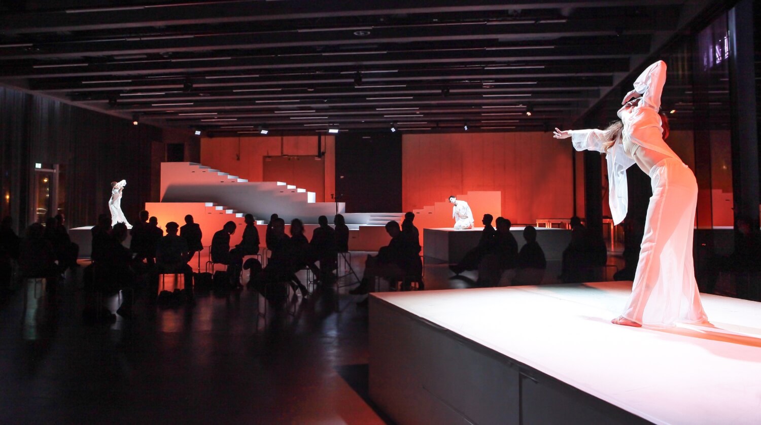 Szene aus Stefano Giannettis Choreografie „Toccata 20“. In der Mitte im Dunkeln sitzendes Publikum. Vorne rechts eine Tänzerin ganz in Weiß, die die Arme ausstreckt zu einer anderen Tänzerin weit hinten links über das Publikum hinweg.