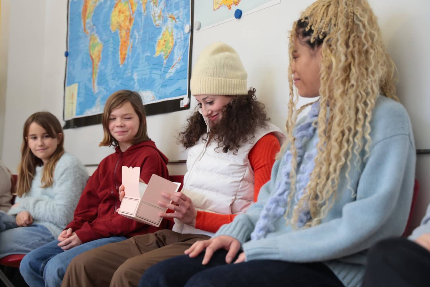 Die beiden Darsteller:innen sitzen am Rand bei den Schüler:innen. Eine schaut lächelnd in eine Papierkiste auf ihrem Schoß.