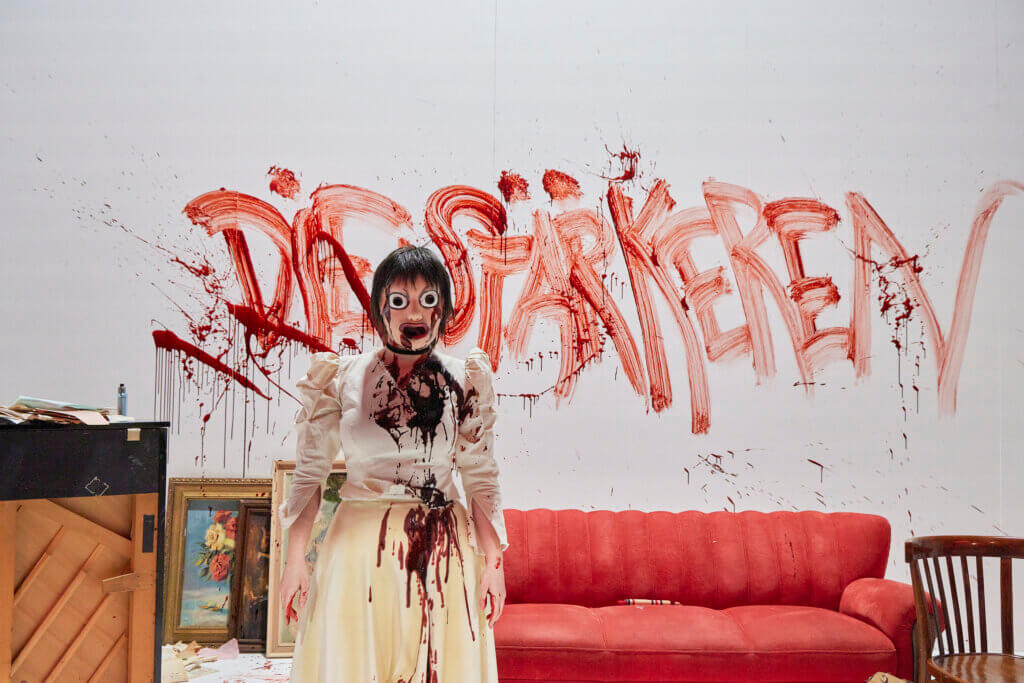 eine Schauspielerin mit Maske, blutverschmiert, dahinter an der Wand ebenfalls mit Kunstblut der Schriftzug „Die Stärkere“