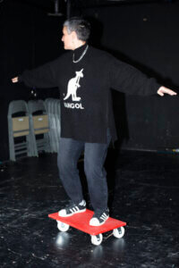 Maya Arad Yasur im Münchner Volkstheater auf einem roten, rollenden Transportbrett, in Skateboardpose.