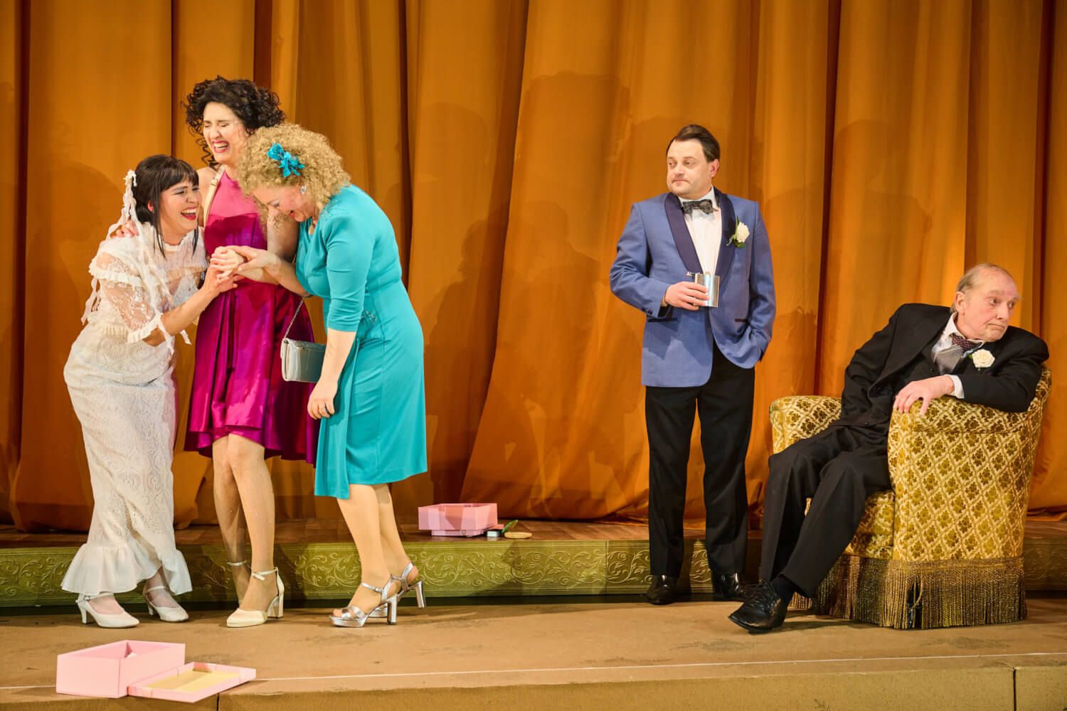 Aufführungsfoto von „Wenn die Sterne fallen“ von Beth Steel am Theater Osnabrück. Ein älterer Mann sitzt rechts in einem Sessel und blickt weg. Ein jüngerer Mann steht neben ihm und schaut auf die drei Frauen, die links stehen und einander zugewandt lachen.