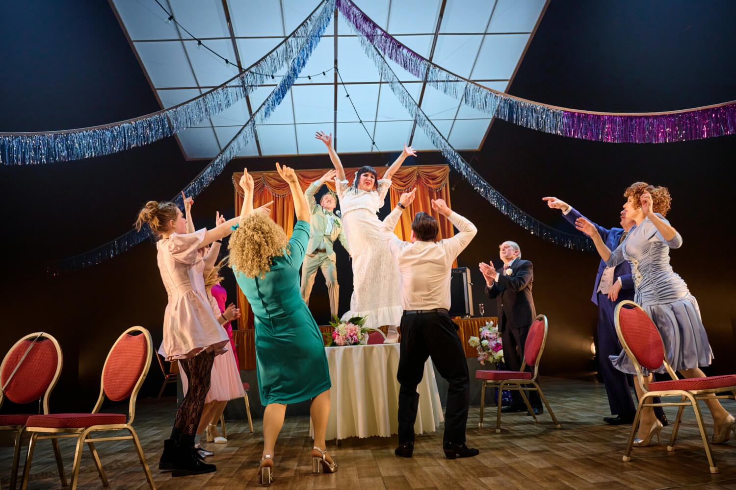 Aufführungsfoto von „Wenn die Sterne fallen“ von Beth Steel am Theater Osnabrück. Eine Hochzeitsgesellschaft tanzt um einen Tisch herum, auf dem Die Braut steht und tanzt.