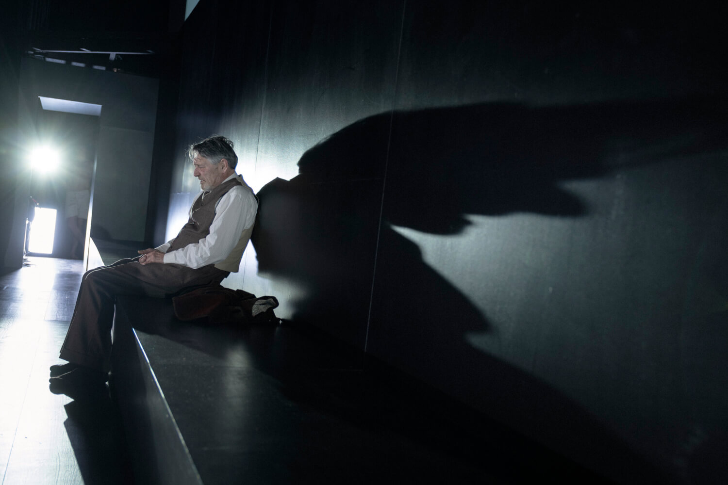 Aufführungsfoto von „Leben und Schicksal“ nach Wassili Grossman, in der Inszenierung von Johan Simons am Schauspielhaus Bochum. Ein Mann sitzt in einem dunklen Gang, der von hinten durch einen Scheinwerfer angeleuchtet wird. Er wirft einen langen Schatten auf die Wand hinter ihm.