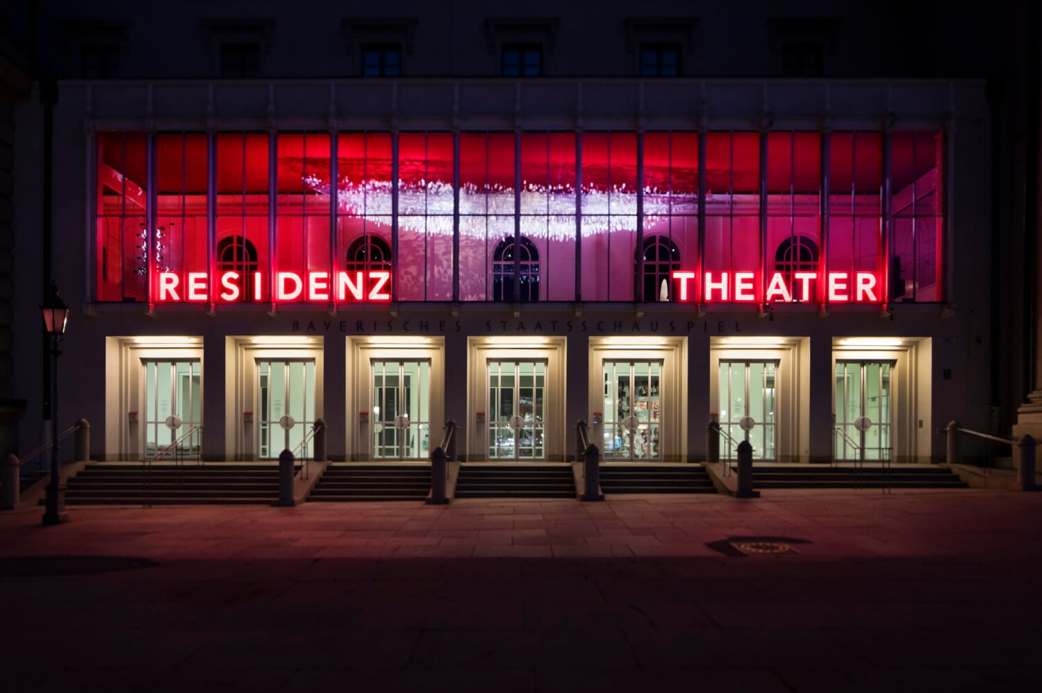Zu sehen ist die Front des Residenztheaters in München. Die sieben Eingangstüren sind hell erleuchtet. Obendrüber auf der zweiten Ebene leuchten groß in rot die Lettern "Residenztheater".