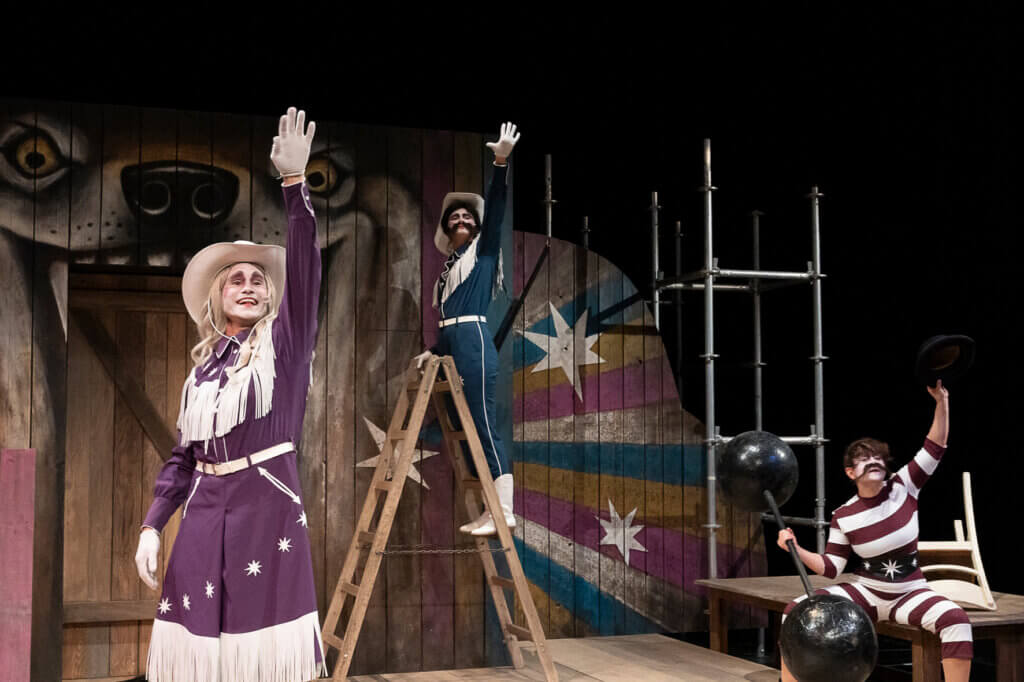 Zwei in Cowboy-Kleidung angezogene Personen heben die rechte Hand zum Himmel. Im Hintergrund steht eine bunt bemalte Holzwand.