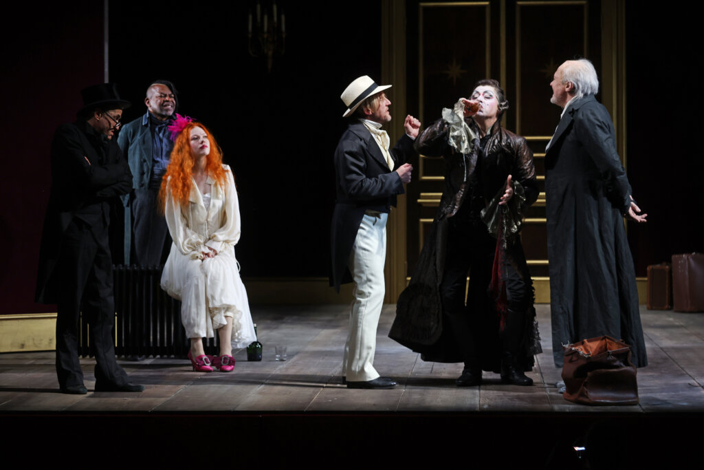 Das Ensemble steht in zwei Grüppchen beisammen, unterhält sich und trinkt.