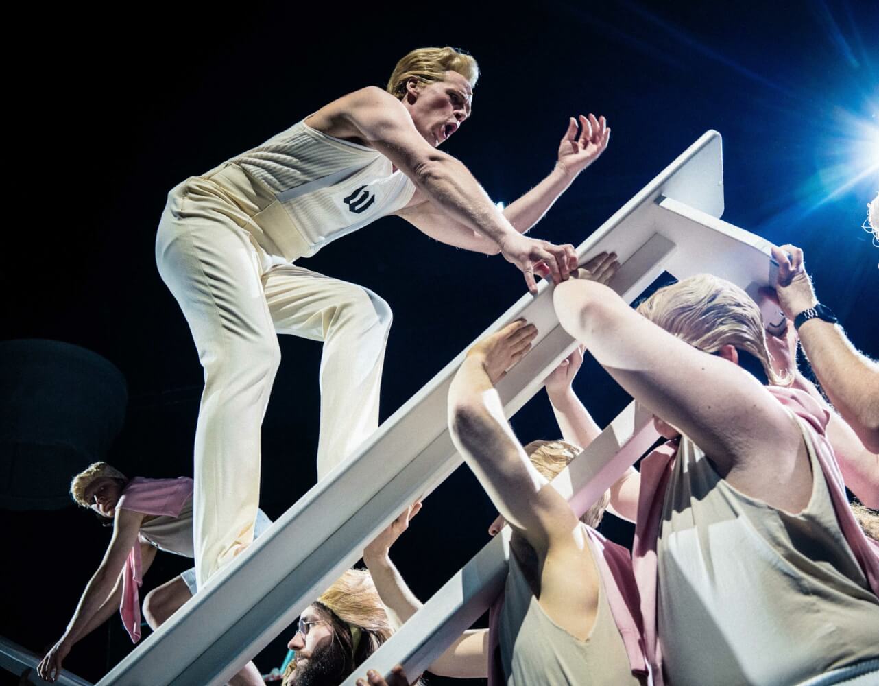 Aufführungsfoto von „Zornfried“ von Philipp Krebs nach dem Roman von Jörg-Uwe Albig am Staatstheater Kassel. Ein Mann mit weißer Hose und weißen Shirt steht auf einer schrägen weißen Bierbank und wird von anderen Menschen in weißen Shirts in die Höhe gehoben.
