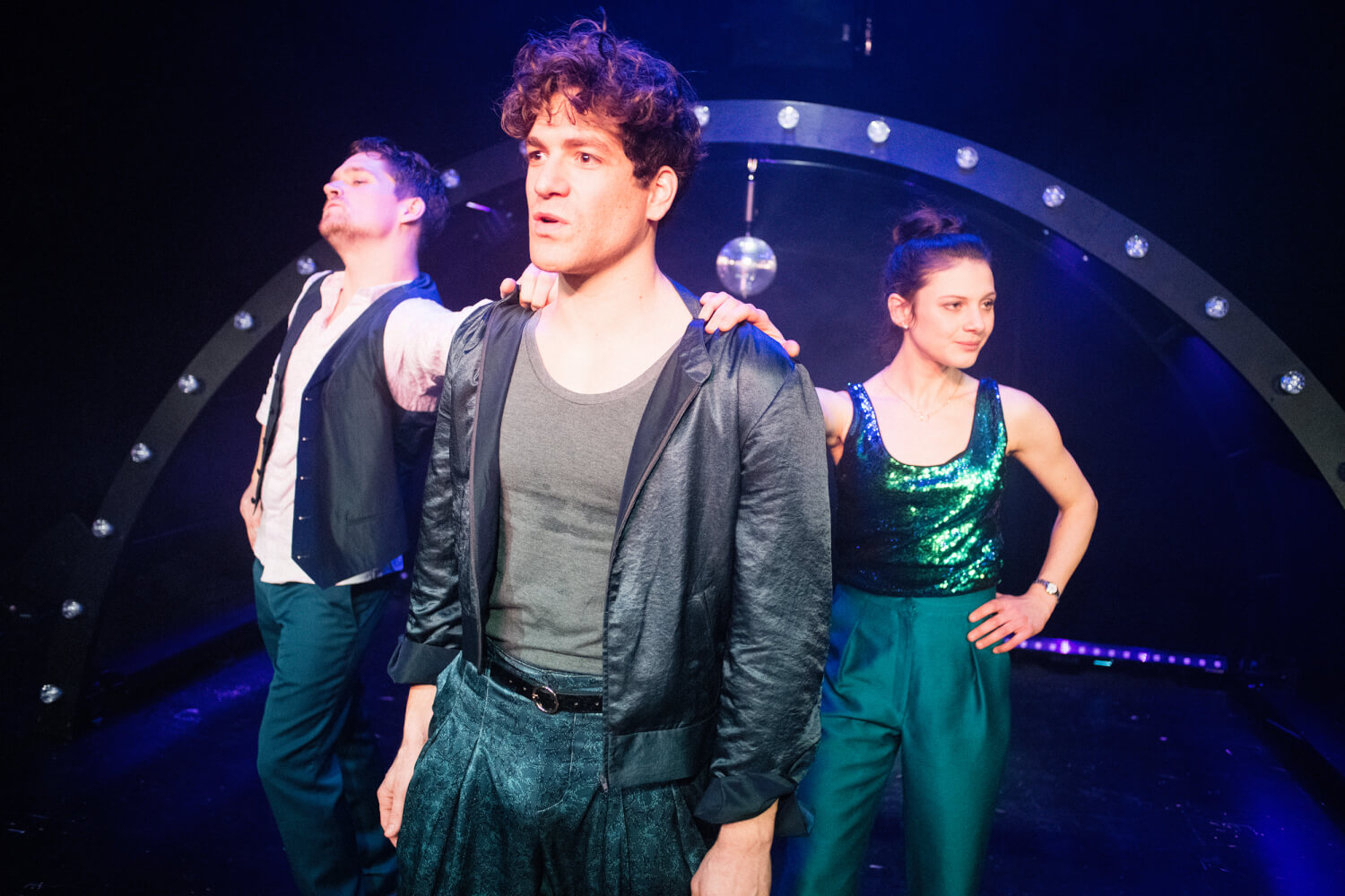 Aufführungsfoto von „Amsterdam“ am Münchner Volkstheater. Ein Mann steht im Vordergrund. Ein Mann und eine Frau stehen seitlich hinter ihm und halten ihn an den Schultern fest. Dahinter ein Halbrund mit Glühbirnen und einer Diskokugel in der Mitte.