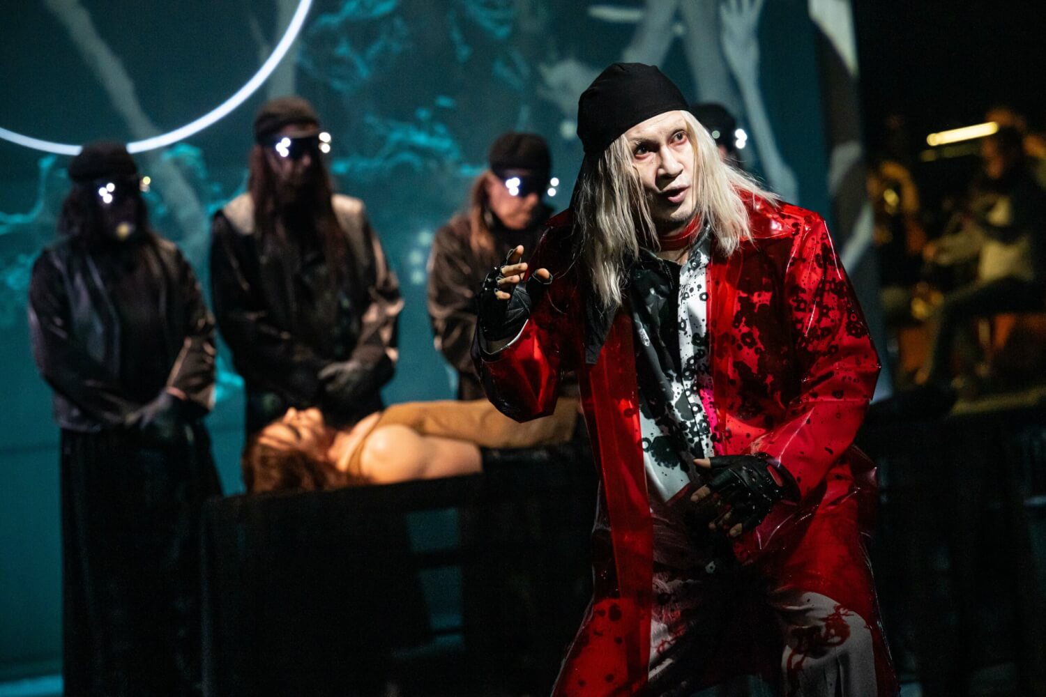 Aufführungsfoto von „Das Spielwerk und die Prinzessin“ von Franz Schreker an den Bühnen Halle. Ein Mann mit rotem Mantel, im Hintergrund drei Männer in schwarz, die hinter einem liegenden Körper stehen.