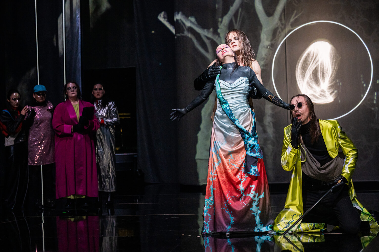 Aufführungsfoto von „Das Spielwerk und die Prinzessin“ von Franz Schreker an den Bühnen Halle. Ein Mann mit dunkler Sonnenbrille und gelbem Mantel kniet mit einem Schwert in der Hand. Daneben hält ein Mann eine Frau fest, die ihre Arme ausbreitet.
