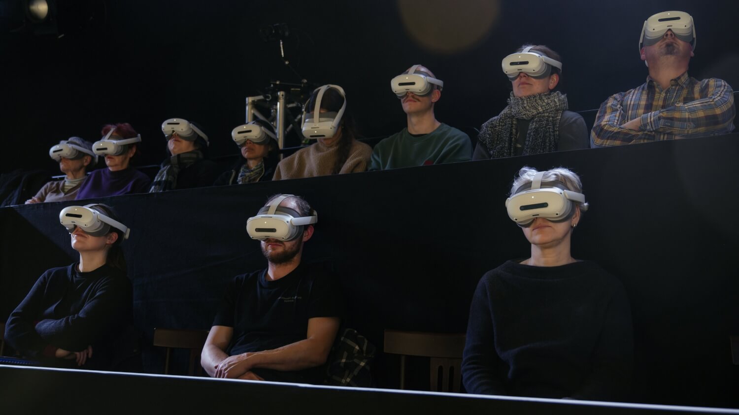 Zuschauer:innen sitzen mit weißen MR-Brillen auf ihren Köpfen in zwei Reihen im Zuschauerraum.