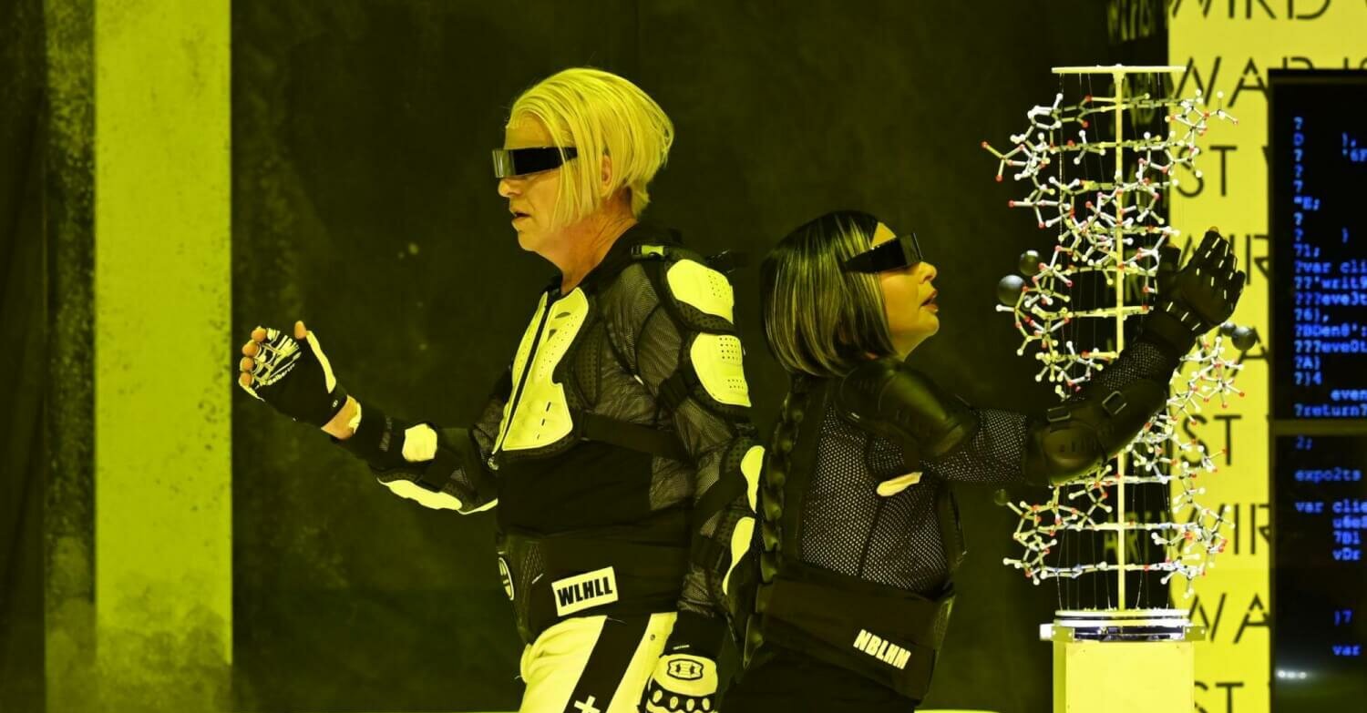 Tilmann Unger und Susanne Serfling stehen Rücken an Rücken. Beide tragen futuristisch aussehende Sonnenbrillen und Anzüge.