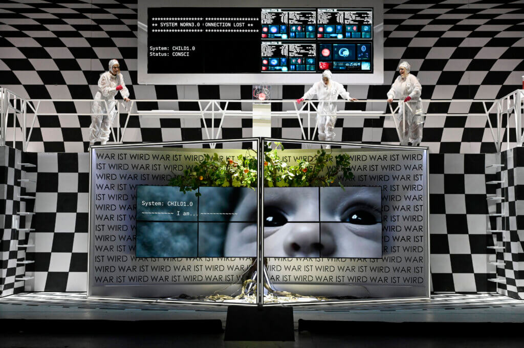 Die Bühne ist in zwei übereinander gelegte Ebenen geteilt. unten ist ein großer Raum mit Bildschirm und diversen Kabeln auf dem Boden. Oben stehen drei Darstellende über eine Brüstung gelehnt. Hinter ihnen ein weiterer Bildschirm mit Code und unterschiedlichen Anzeigen.