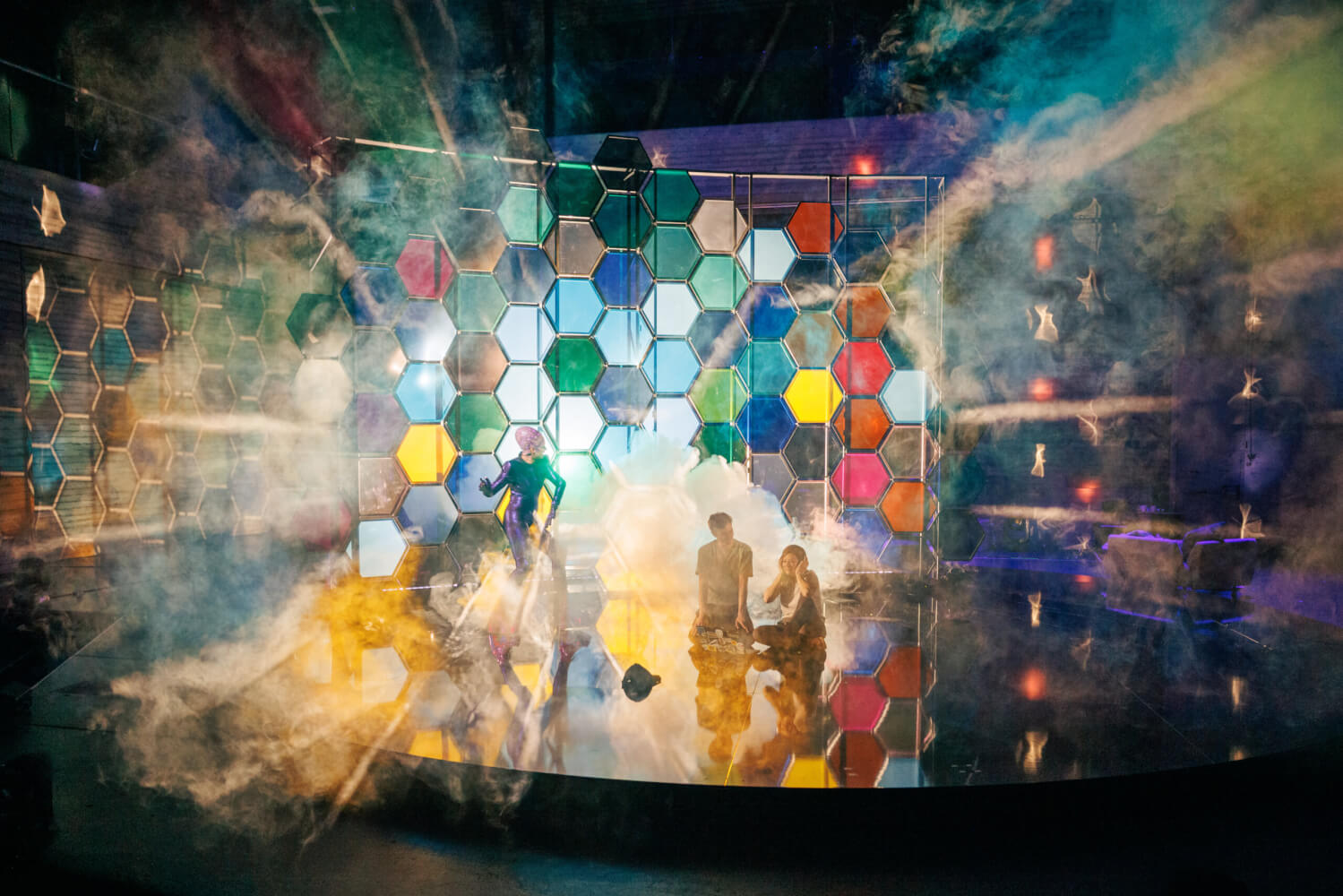 Aufführungsfoto von „Moskitos“ von Lucy Kirkwood am Badischen Staatstheater Karlsruhe. Ein knallbuntes wabenartiges Bühnenbild, aus dem nebeliges Licht strahlt. Davor steht eine Frau und ein Mann und eine Frau sitzen auf der Bühne.