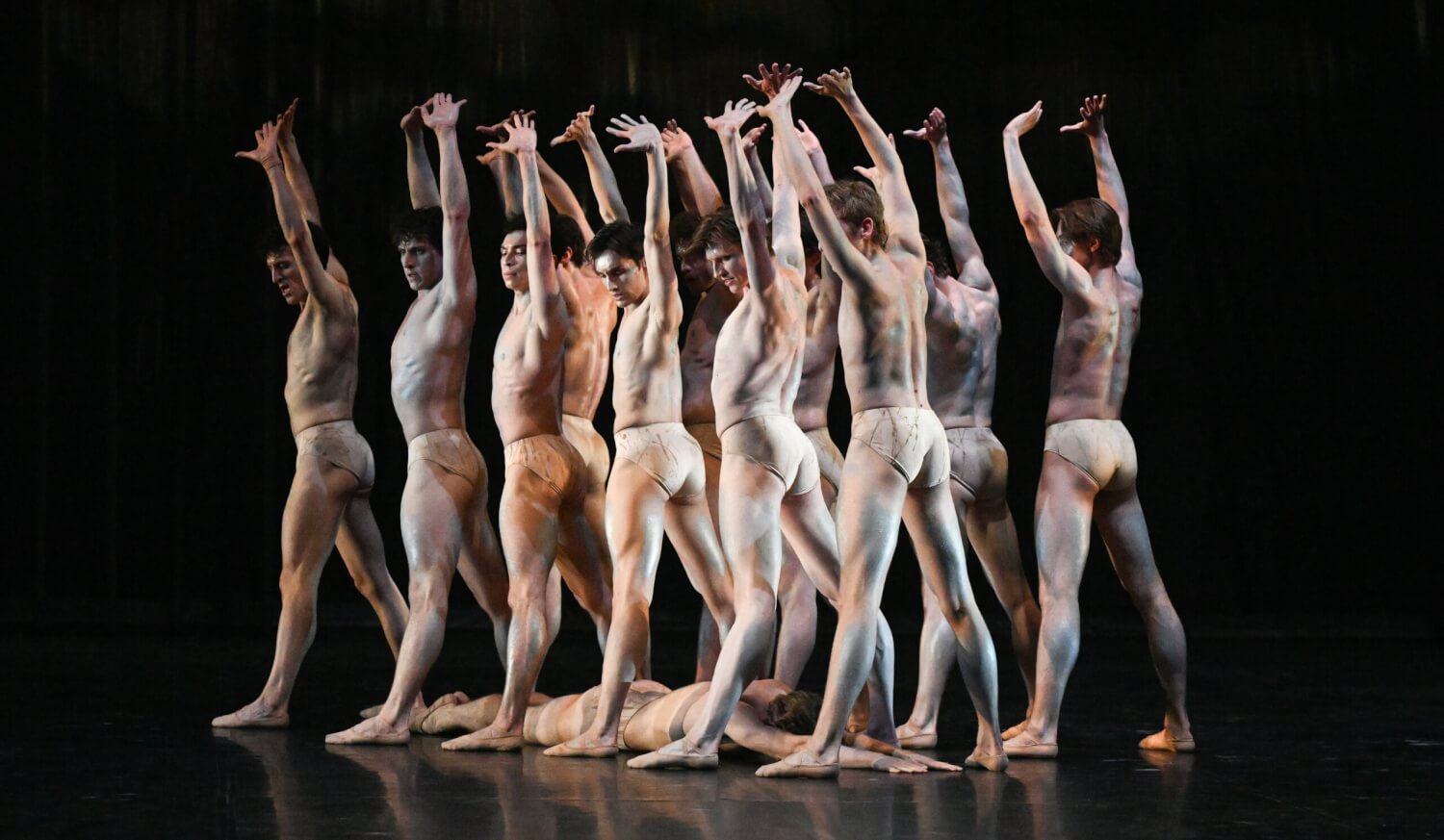 „Le Sacre du Printemps“ aus „Tribute to Tetley“ Glen Tetley des Stuttgarter Ballett. Das männliche Tänzerensemble streckt die Arme nach oben und steht breitbeinig hinterainder. Unter ihnen liegt ein Tänzer bäuchlinks auf dem Boden.