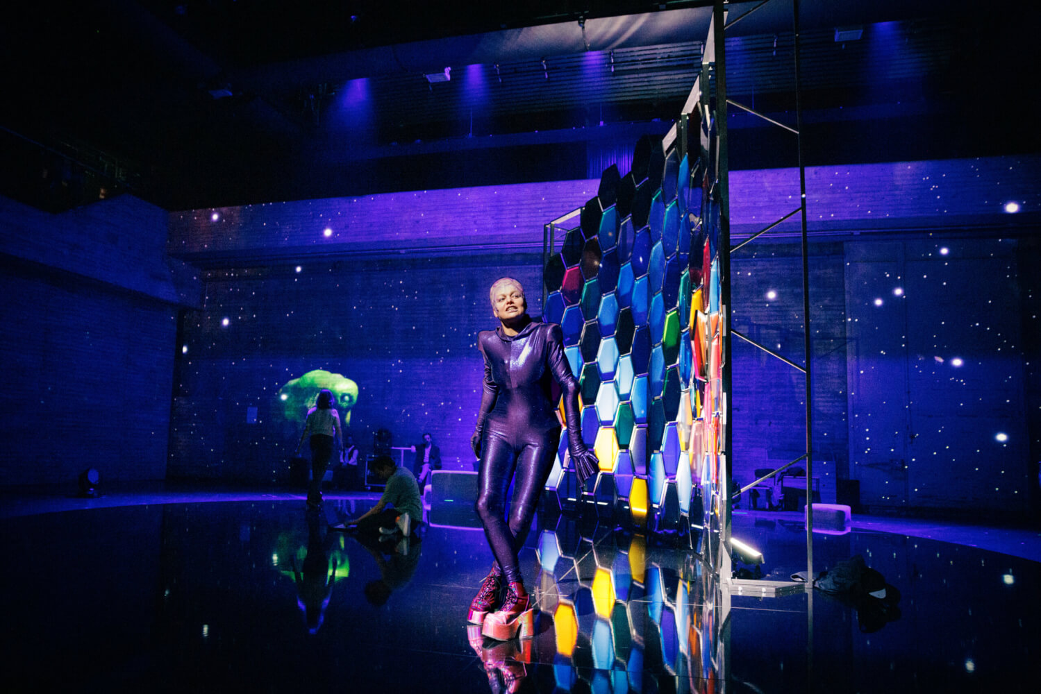 Aufführungsfoto von „Moskitos“ von Lucy Kirkwood am Badischen Staatstheater Karlsruhe. Vor einem Sternenhimmel steht schräg eine bunte Wabenwand. Davor ist eine Frau in Plateauschuhen und hautengem Ganzkörperanzug.