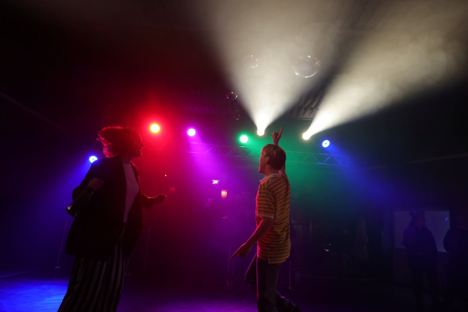 Aufführungsfoto von „Düsterbusch City Lights“ nach dem Roman von Alexander Kühne an der Neuen Bühne Senftenberg mit Richard Fuchs und Lene Juretzka. Eine Frau und ein Mann tanzen in einem dunklen Raum, der mit bunten Scheinwerfern beleuchtet ist.