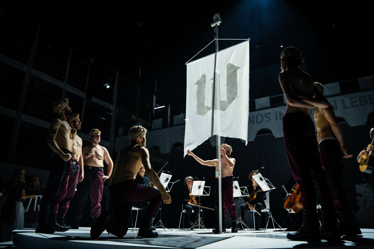 Aufführungsfoto von „Zornfried“ von Philipp Krebs am Staatstheater Kassel. In der Mitte ein Fahnenmast mit einer weißen Fahne und einem schwarzen W. Drumherum oberkörperfreie Menschen und Musiker im Hintergrund.
