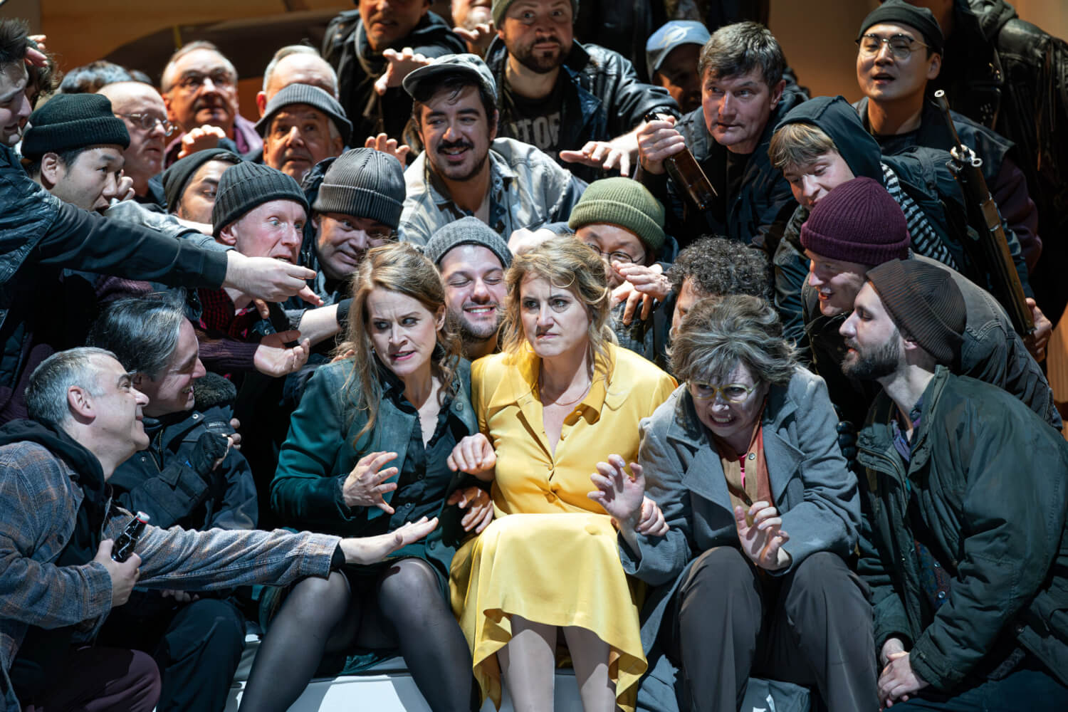 Aufführungsfoto von „Regina“ von Albert Lortzing an der Oper Leipzig. Eine Frau im gelben Kleid sitzt in der Mitte einer dunkel gekleideten Menschenmasse, die sich zu ihr lehnen und die Arme nach ihr ausstrecken.