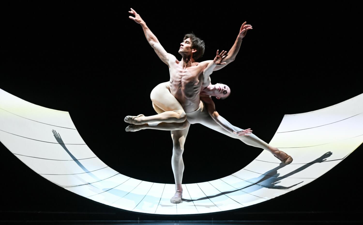 „Ricercare“ aus „Tribute to Tetley“ Glen Tetley des Stuttgarter Ballett. In einem Halbrund steht ein Tänzer aufgedehnt, eine Tänzerin schlingt sich um seinen Oberkörper.