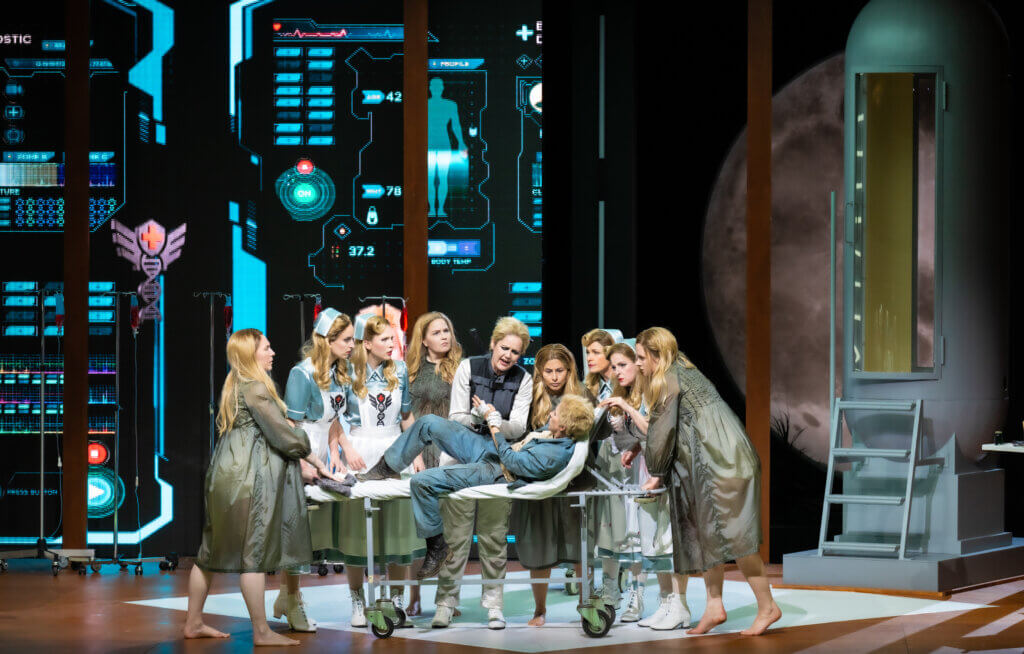 Vor aufleuchtenden Bildschirmen an der Bühnenwand steht das Ensemble um ein Krankenbett versammelt. Darauf liegt Astrid Kessler und schaut Trine Trine Møller in die Augen, die bewegt ihre Hand hält.