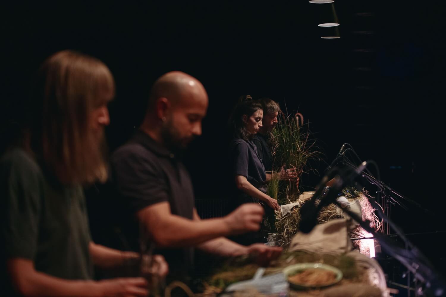 Aufführungsfoto von „Cow | Deer“ von Katie Mitchell, Nina Segal und Melanie Wilson an der Schaubühne Berlin. Vier Menschen stehen nebeneinander aufgereiht an einem langen Tisch mit Mikrofonen, auf dem sich viele Gegenstände zur Lauterzeugung befinden.