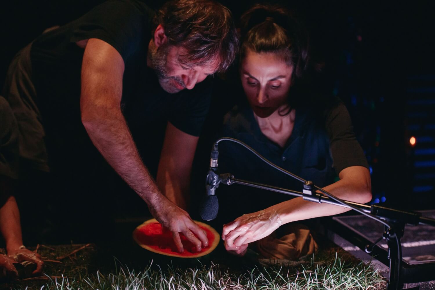 Aufführungsfoto von „Cow | Deer“ von Katie Mitchell, Nina Segal und Melanie Wilson an der Schaubühne Berlin. Ein Mann und eine Frau beugen sich über ein Mikrofon und eine halbe Wassermelone, die auf Gras liegt. Der Mann fasst mit seinen Fingern in die Melone.