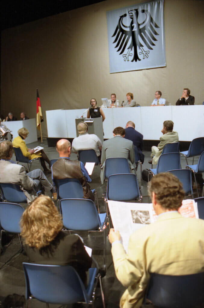 Experten des Alltags in „Deutschland 2“. Menschen sitzen im Halbkreis auf Stühlen und wohnen einer Pressekonferenz bei.