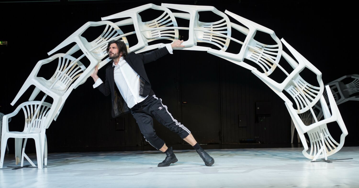 Giuseppe Spota bei unserem Covershooting im Bühnenbild zu seiner Choreographie „Momo“ am Musiktheater im Revier in Gelsenkirchen. In einem Bogen aus weißen Plastikgartenstühlen steht Spota schräg nach links geneigt und hält isch mit beiden Händen an Stuhlbeinen fest.