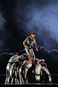 Ensembleszene aus Spotas Choreographie „Tambora“ am Staatstheater Mainz. Menschen im mit silbernen Anzügen heben eine Frau im silbernen Anzug in die Höhe.