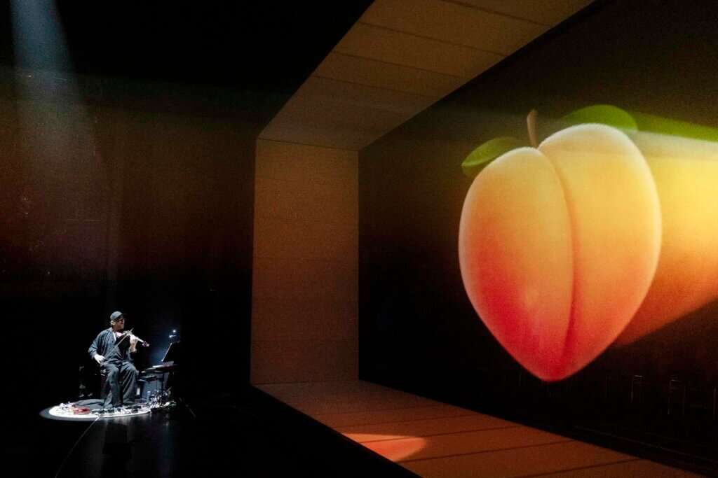Rechts wird ein riesiges Pfirsich-Emoji an die Wand projiziert. Links sitzt Matthias Jakisic und macht Musik.