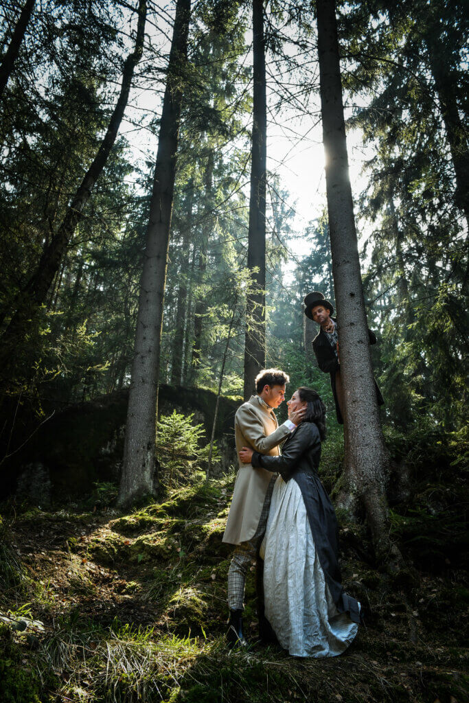 Szene aus der Uraufführungsproduktion von „Zucker“. Ein Mann und eine Frau stehen in einem Wald und umarmen sich. Im Hintergrund schaut ein Mann mit Zylinder hinter einem Baumstamm hervor.