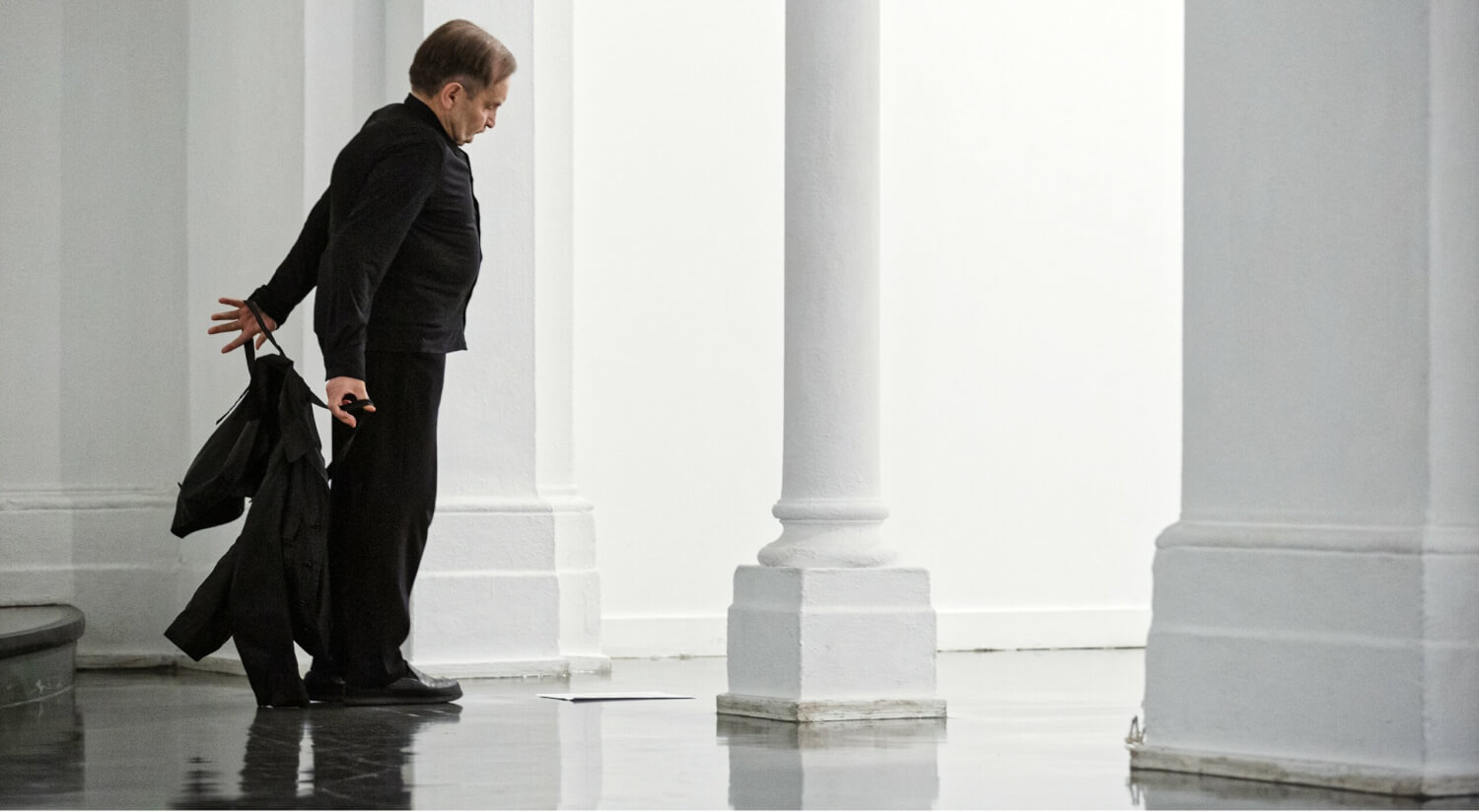 Raimund Hoghe in einem schwarzen Anzug vor weißen Säulen auf der Bühne.