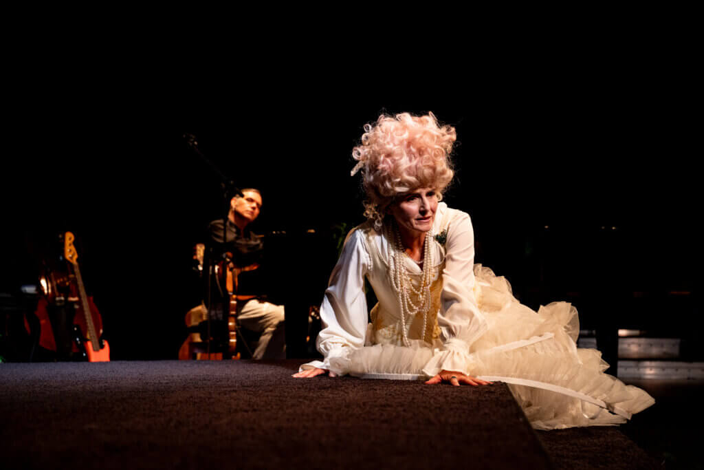 Aufführungsfoto von „Orlando“ am Theater Osnabrück. Eine Frau sitzt eingesunken da, gekleidet in einem weißen Reifrock, Perlenketten und weiß-rose Lockenperücke.