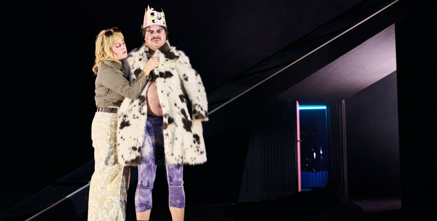 Aufführungsfoto von „Sankt Falstaff“ am Theater Osnabrück. Eine Frau steht neben einem Mann mit Pappkrone, der einen gefleckten Kuhmantel an hat.