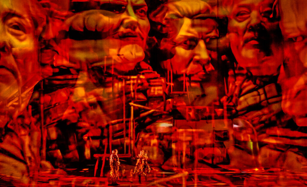 eine rot bestrahlte Bühne mit mehrern Darsteller:innen und Projektionen auf Gaze