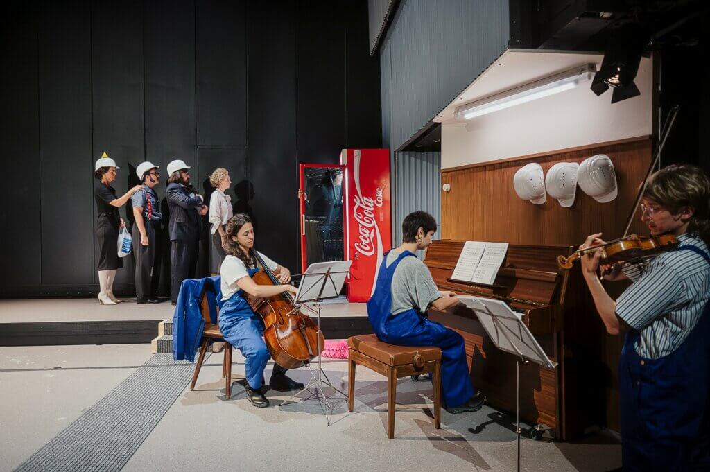Aufführungsfoto von „Stahlgipfel“. Im Hintergrund vier Personen stehen in einer Reihe. Im Vordergrund: Ein Mann spielt eine Geige, eine Frau sitzt am Klavier und eine Frau spielt Cello.