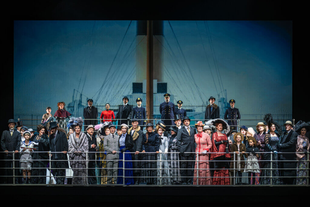 Aufführungsfoto von „Titanic“ am Theater Chemnitz. Ensembleszene am Schiffgeländer, dahinter die Schiffsoffiziere und der Schornstein des Schiffes.