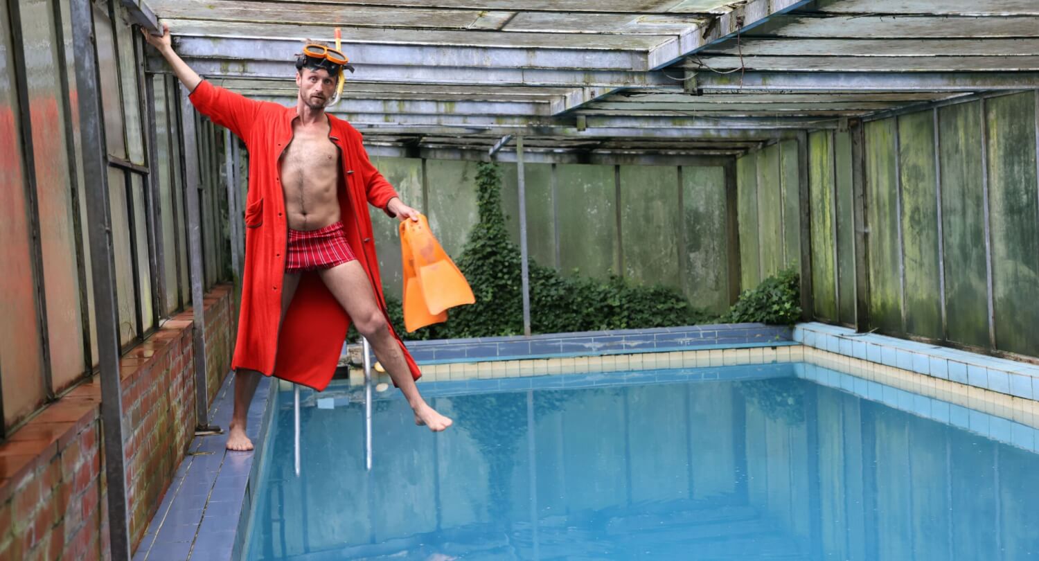 Werbefoto zu Samir Akikas Produktion „(Little) Mr. Sunshine“. Er steht am Beckenrand eines Pools mit einem roten Bademantel und orangefarbenen Flossen in der Hand.