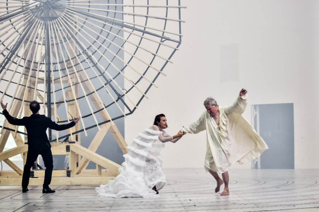 Aufführungsfoto von „Lear“ am Deutschen Theater Berlin 2019. Eine Frau in weißem Gewand zieht einen älteren Mann in einem weißen Gewand über die Bühne.